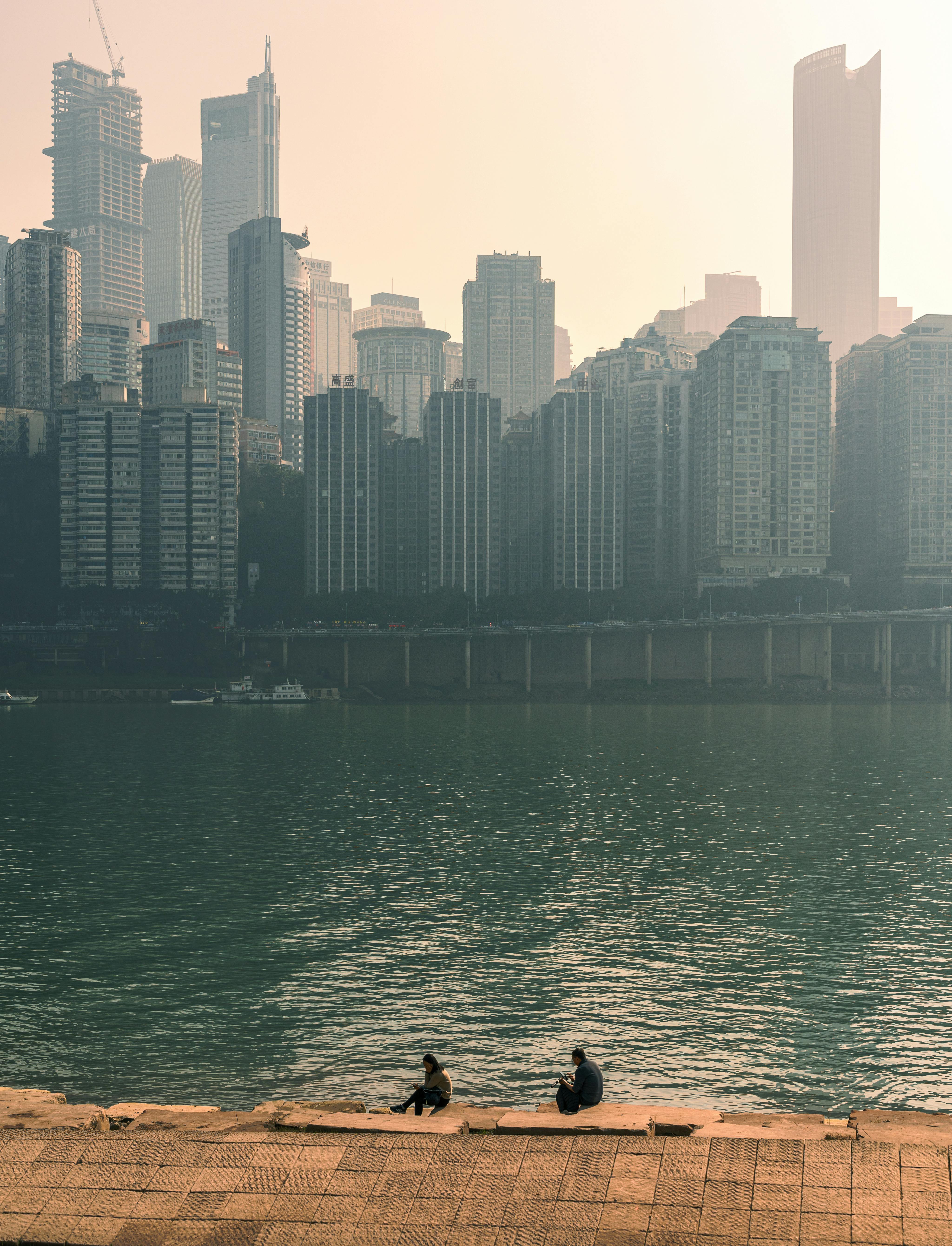 Two people by a river with a modern city skyline in the background on a hazy day.