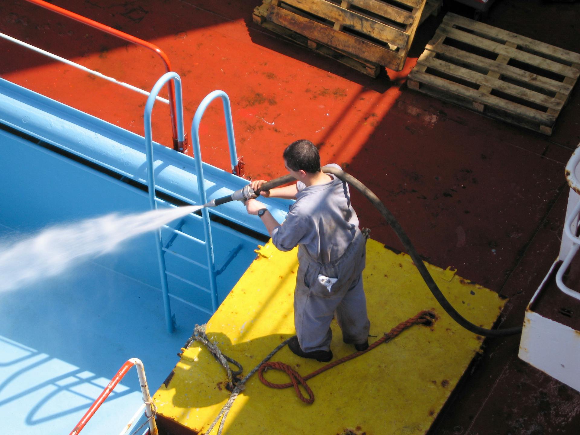 A worker using a high-pressure hose to clean the deck of a ship, showing industrial processes.