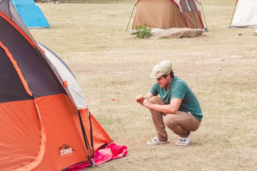 A man in casual attire sets up an orange tent at a campground during the day, showing a relaxed outdoor activity.