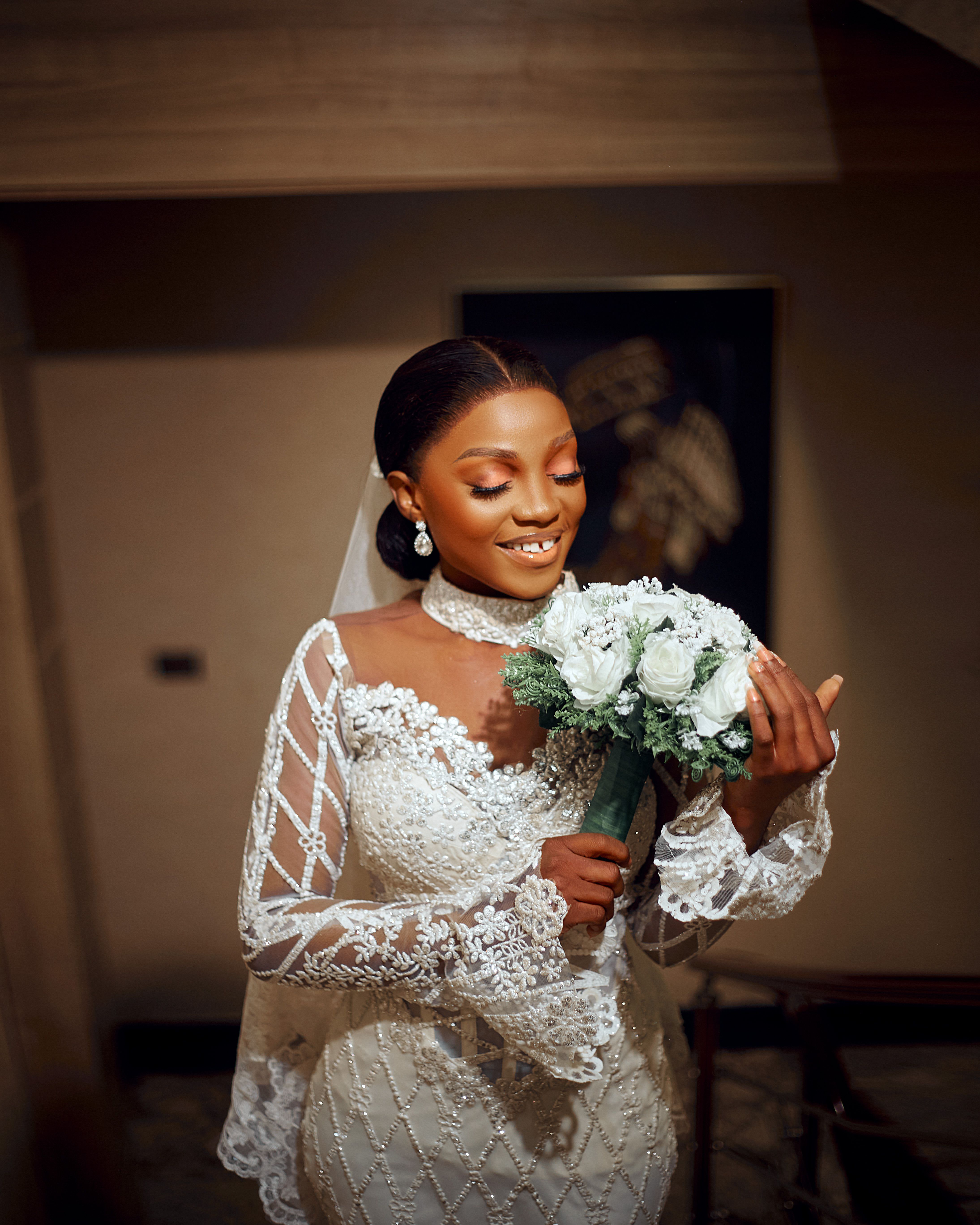 Beautiful bride in lace gown holding a bouquet, radiating joy indoors.
