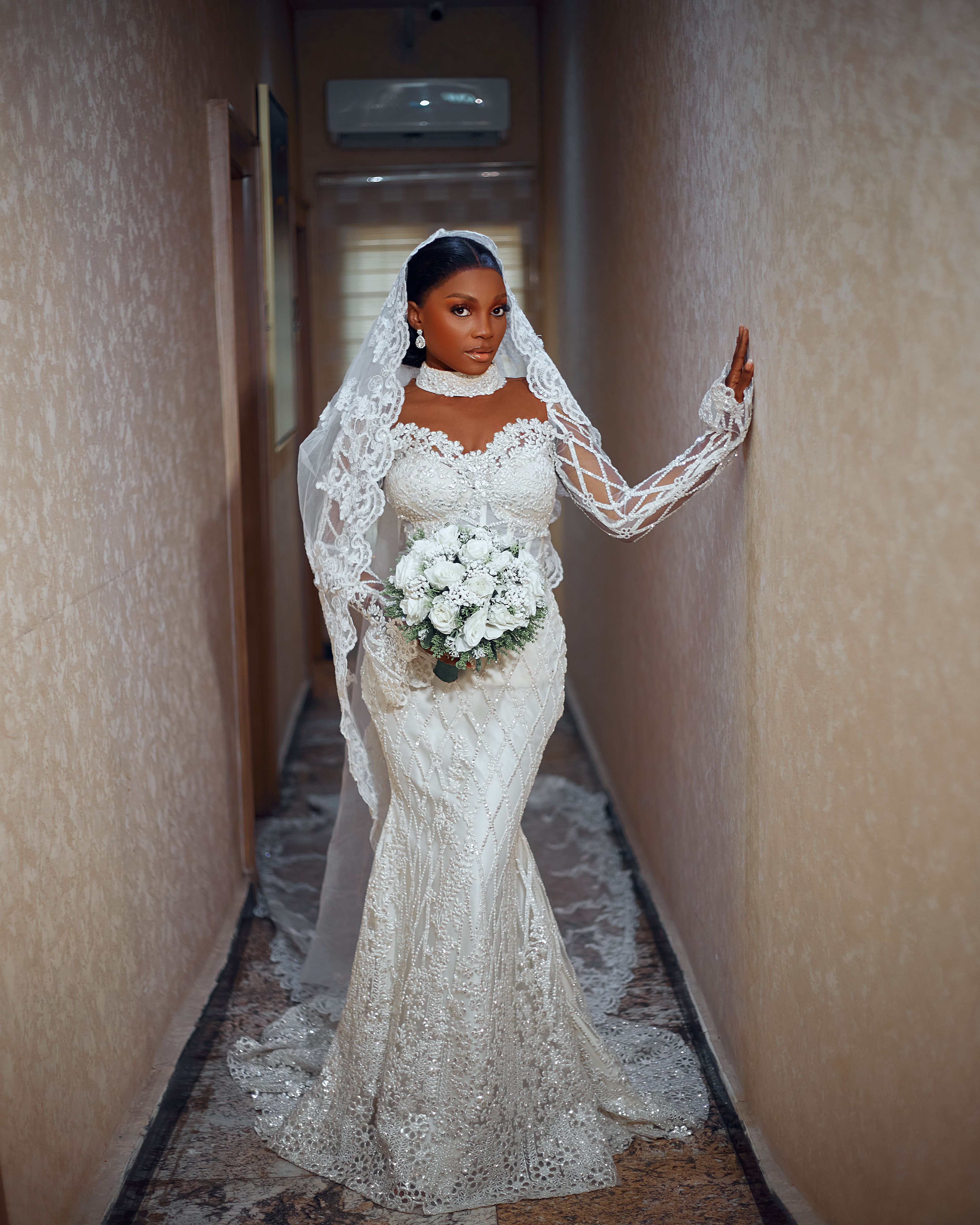 Stunning bride in a detailed lace gown holding a white bouquet, captured in a narrow hallway.