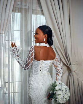 A stunning bride in a lace dress holding a bouquet, looking out a window.