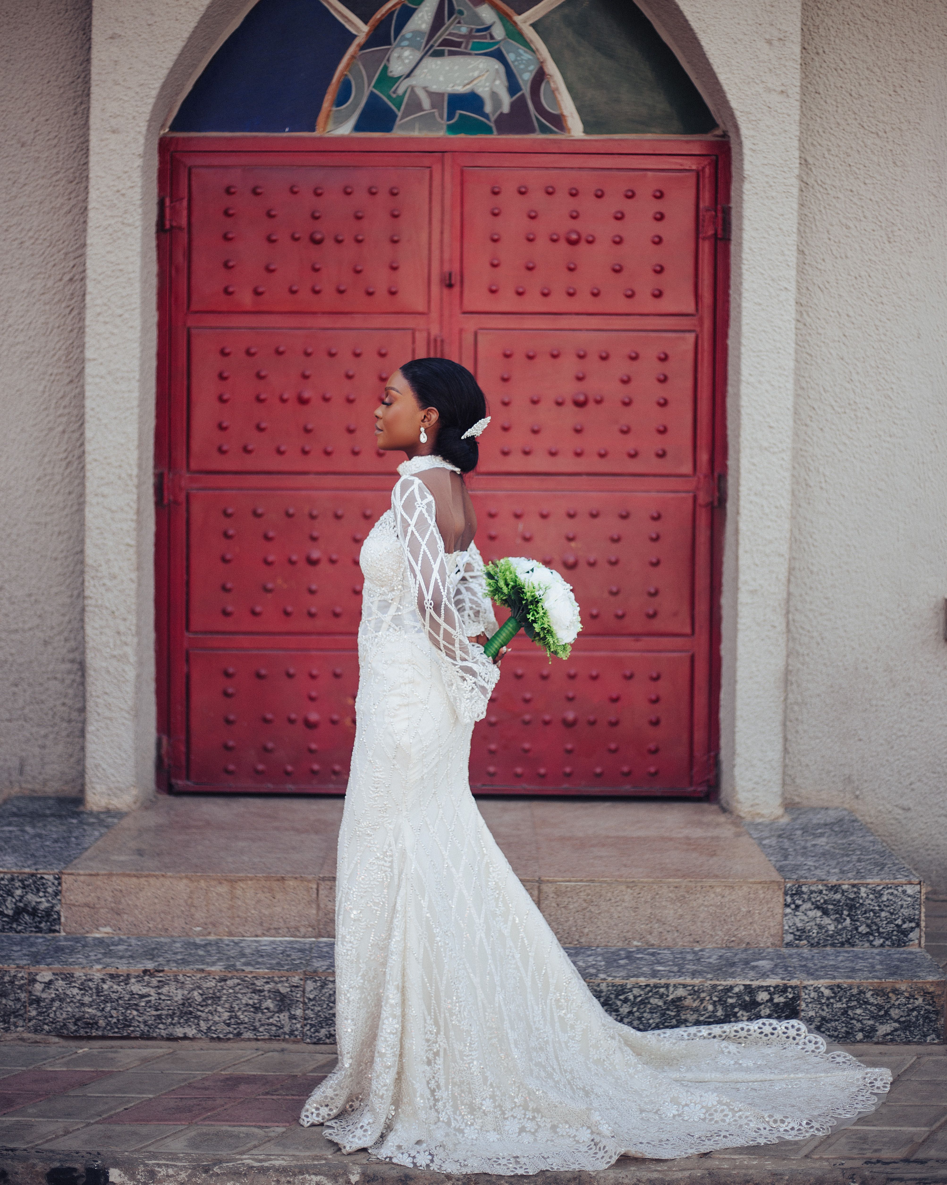 Bride in a lace dress holding flowers stands by a red church door.