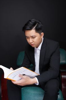 A well-dressed young man in a suit intensely reads a book indoors.