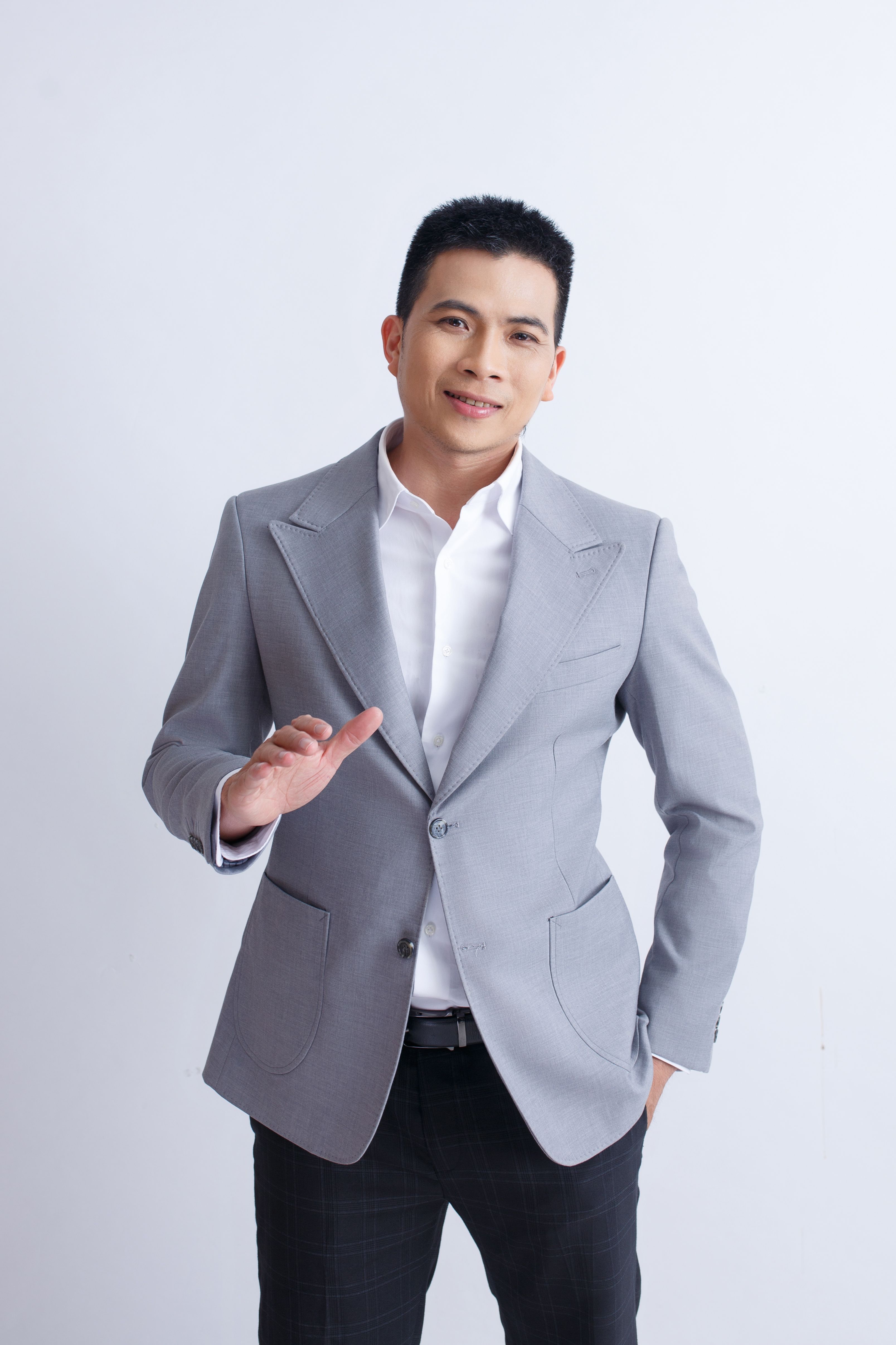 Confident man wearing a grey suit, standing with a welcoming gesture against a white background.