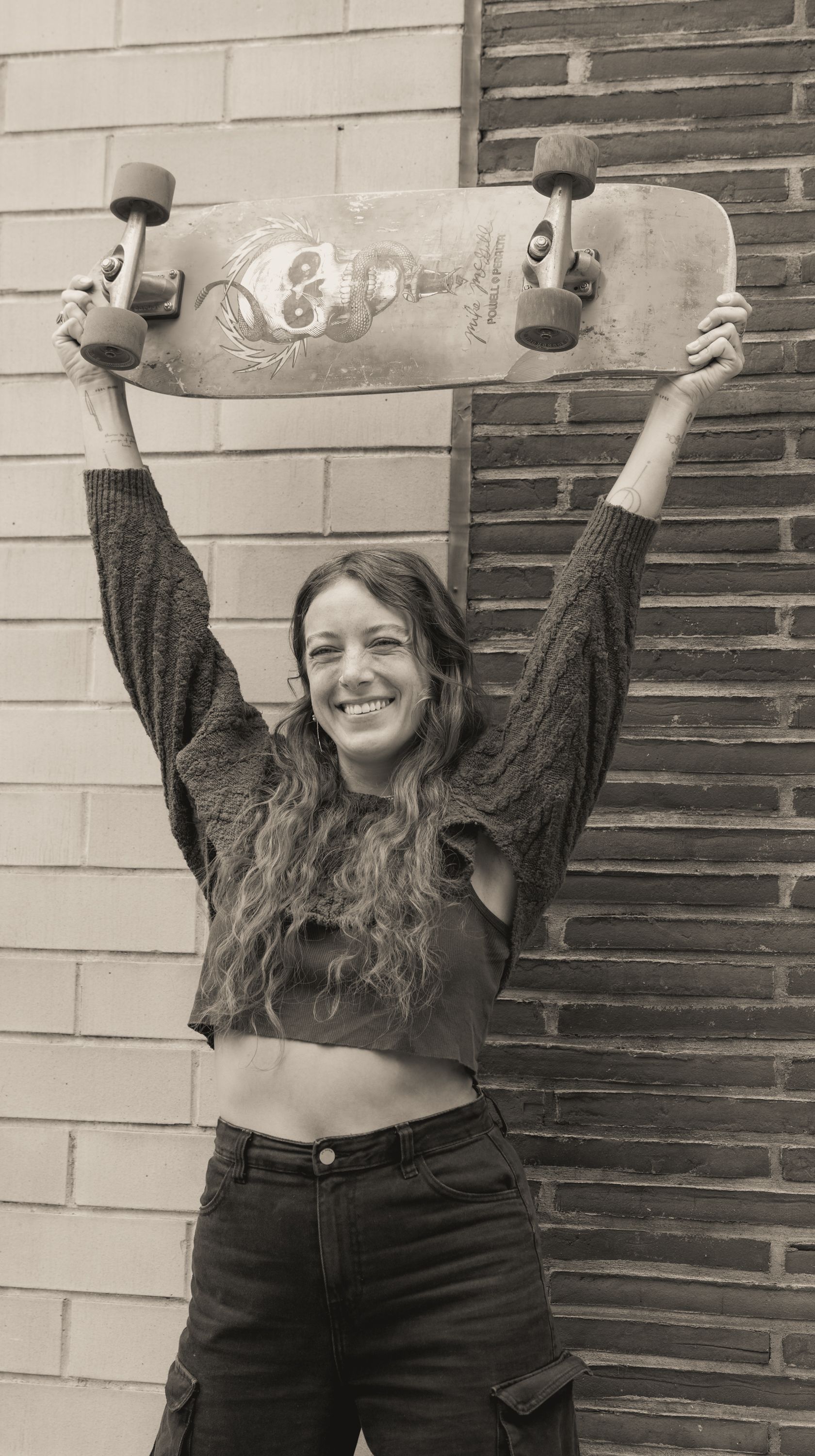 A smiling woman holds up a skateboard against an urban backdrop in New York City.
