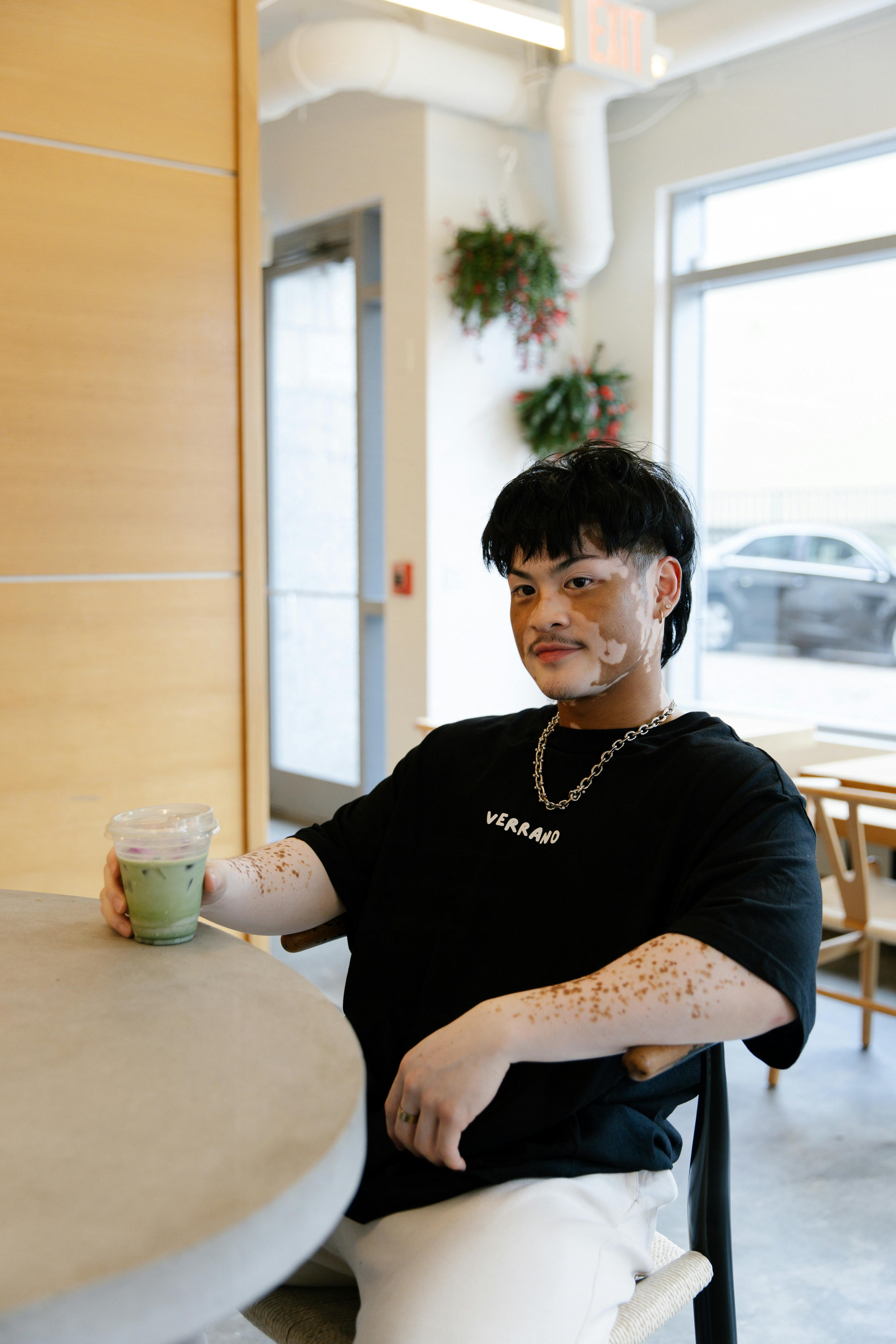 A man with vitiligo sits relaxed at a cafe table, sipping a cold brew coffee in a cozy indoor setting.