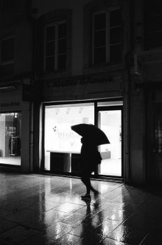 A lone figure with umbrella walks past brightly lit storefront at night.
