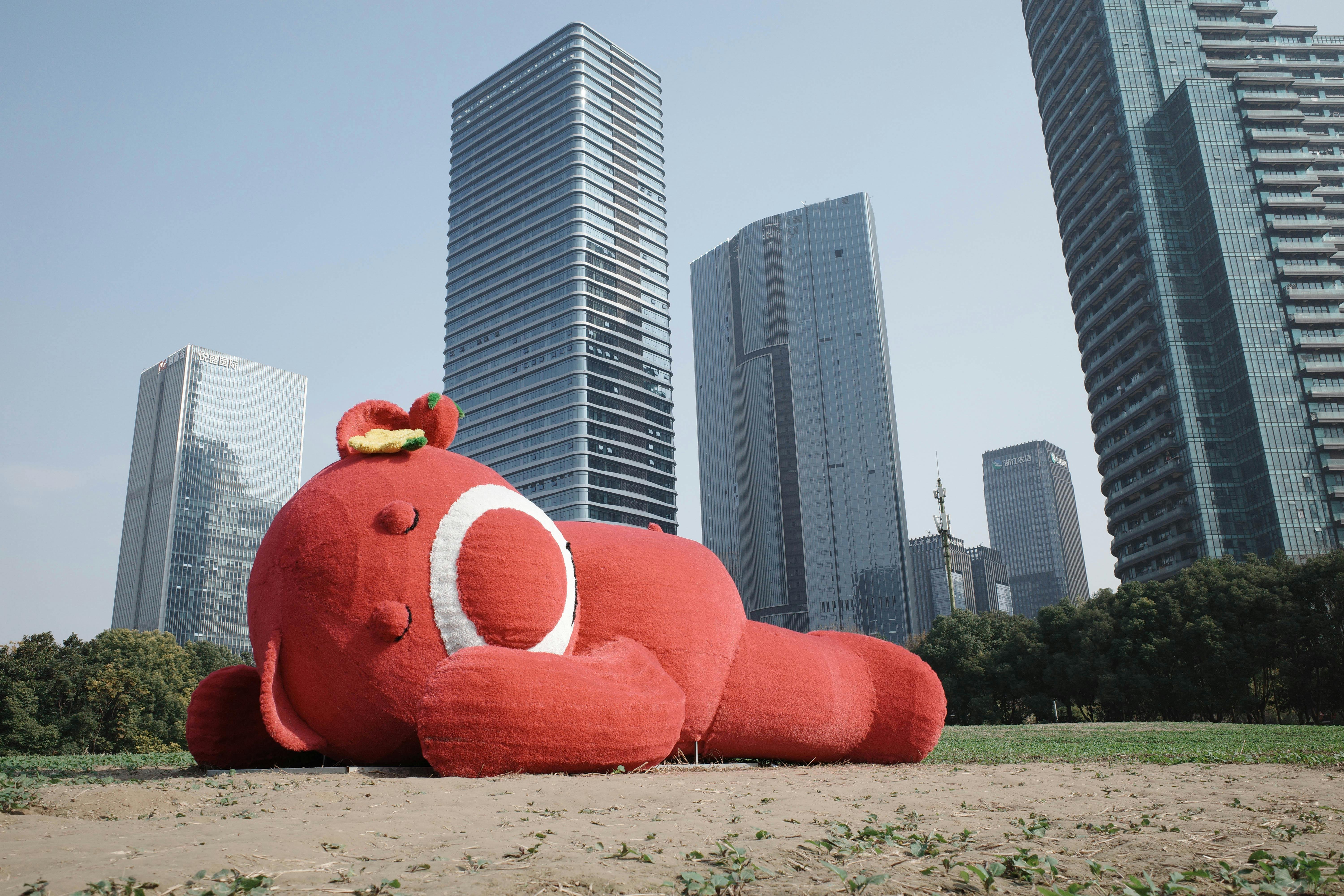 A large red toy sculpture lying on grass with towering skyscrapers in the background under a clear sky.