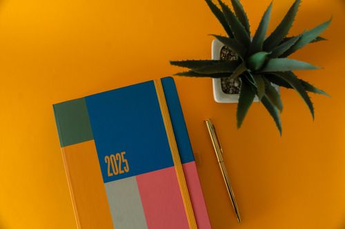 Free Colorful 2025 planner with gold pen and plant on yellow background. Stock Photo