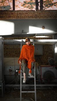 A woman with an orange shawl sits in an industrial setting with brick walls.