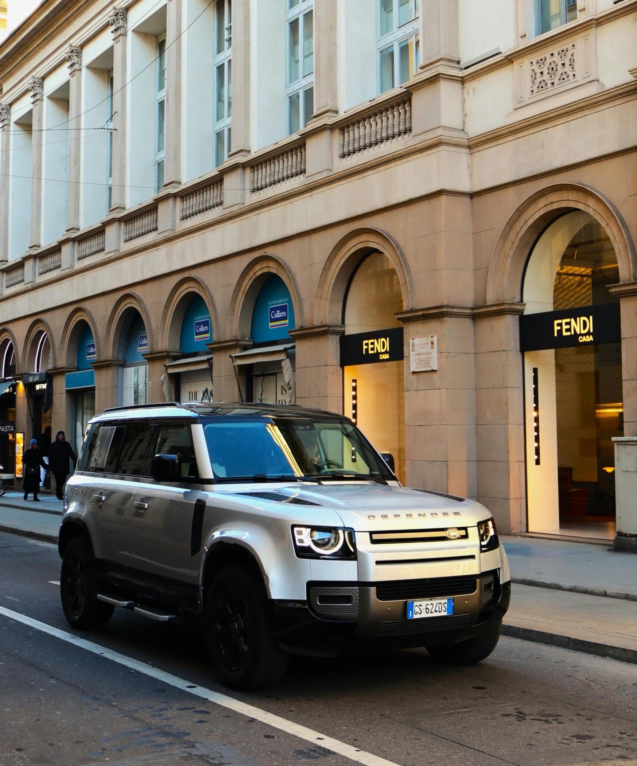 Gratis Sebuah Land Rover Defender berwarna perak diparkir di depan sebuah toko mewah di Milan, Italia. Foto Stok