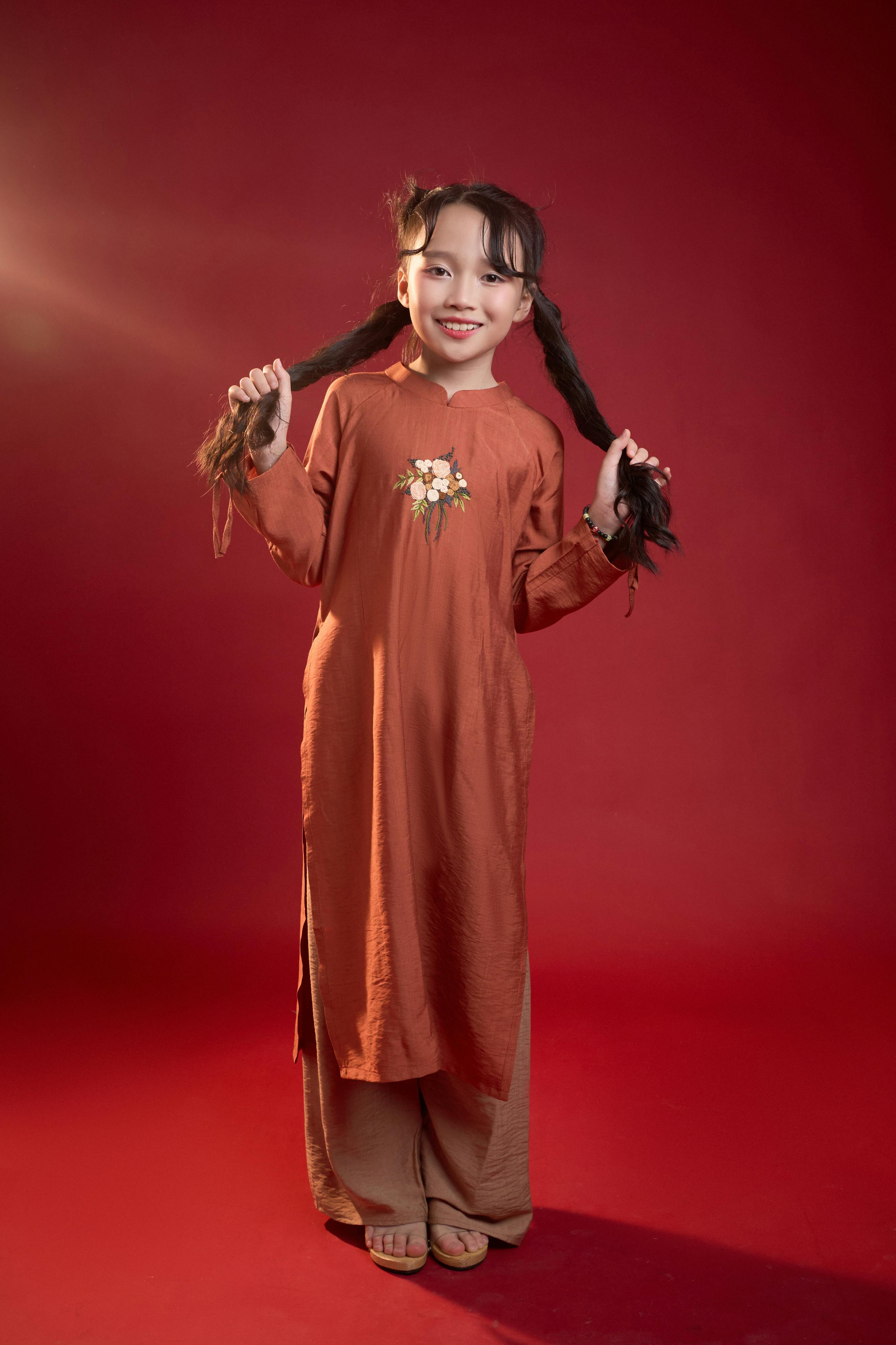 Gratis Ung pige i traditionel ao dai med pigtails poserer mod en levende rød baggrund, der viser vietnamesisk kultur og mode. Lagerfoto
