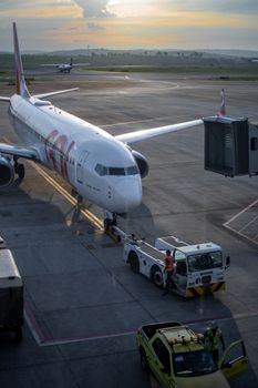 A commercial airplane at the Minas Gerais airport during sunset, with boarding bridge and ground crew visible.