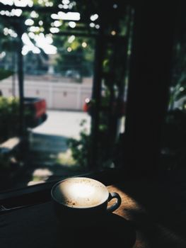 A steaming cup of coffee sits on a table, bathed in natural daylight near a window.