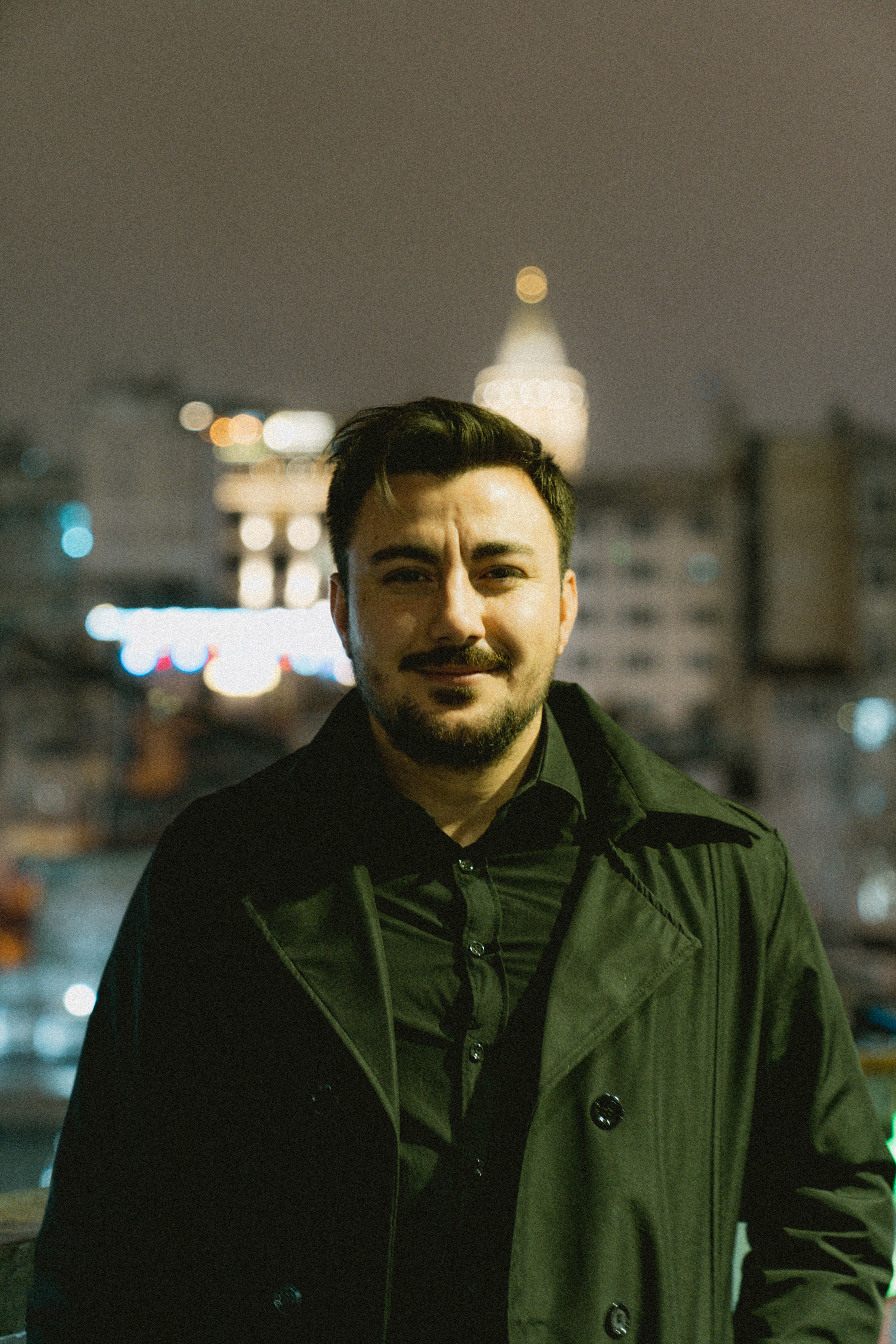 Portrait of a man in a black coat standing in front of a city skyline at night.