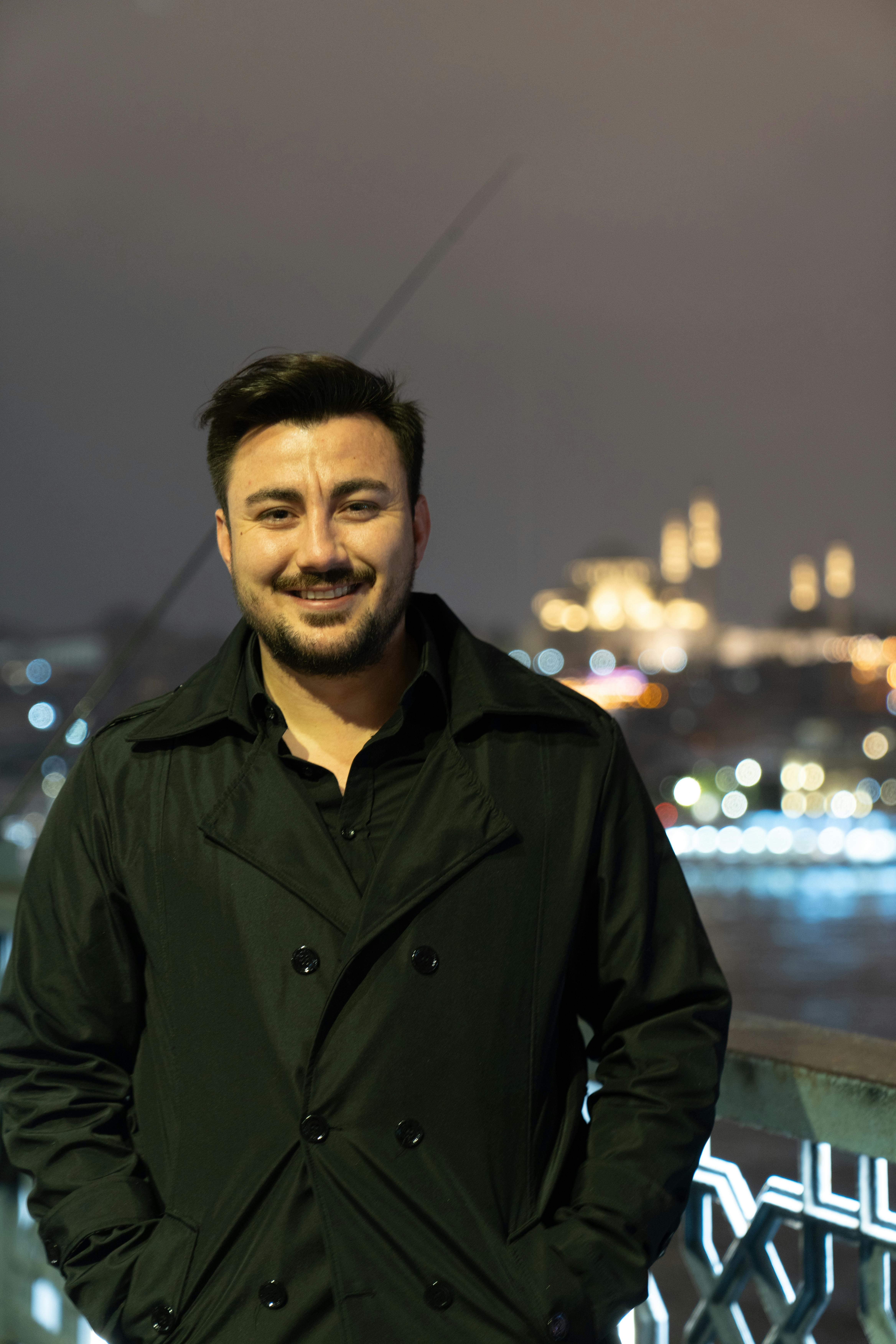 A man in a black coat smiles on a city bridge at night with blurred lights behind.