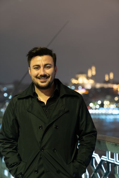 Free A man in a black coat smiles on a city bridge at night with blurred lights behind. Stock Photo