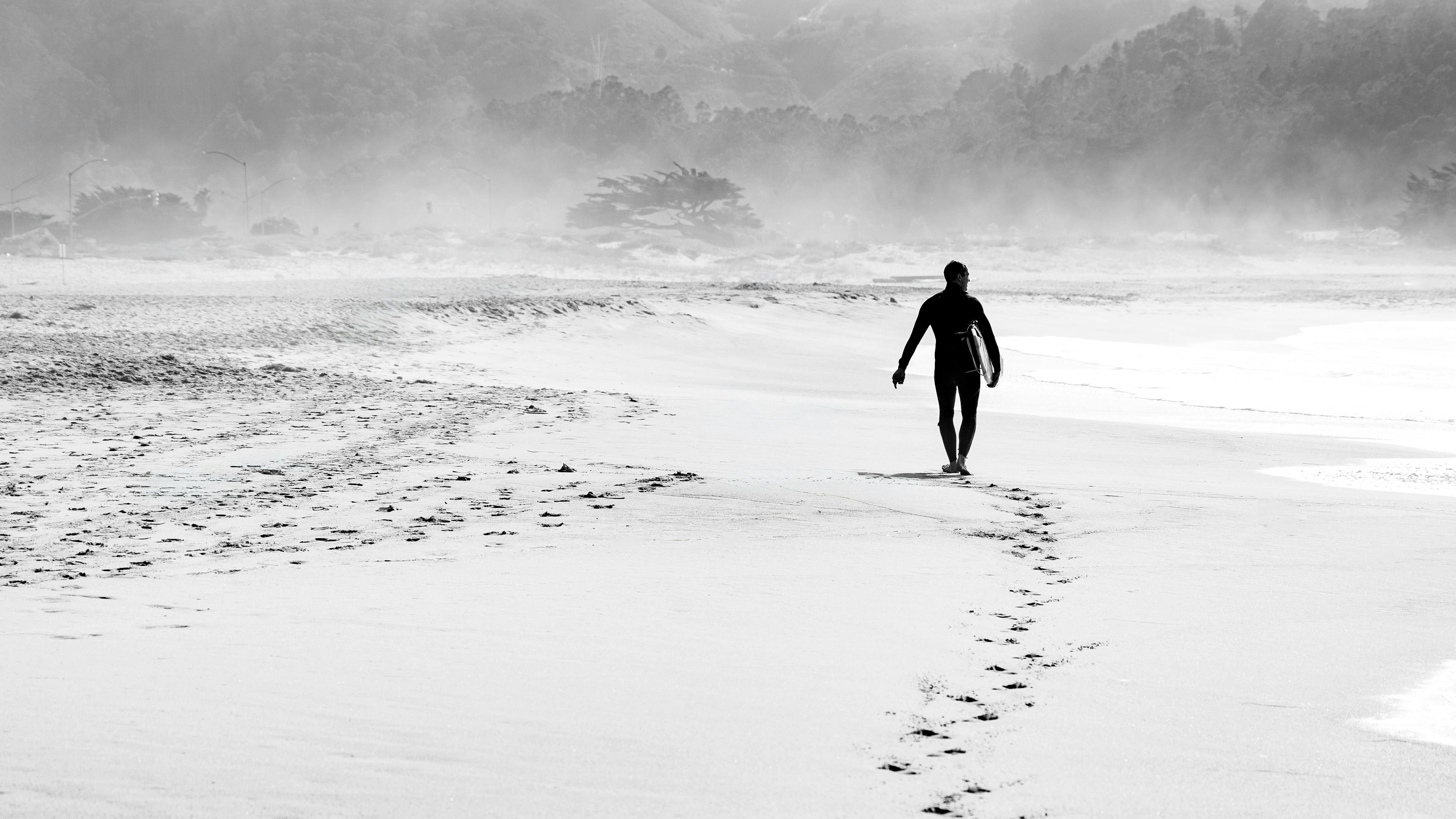 Black and white photo of a surfer walking along a foggy beach, leaving footprints in the sand.