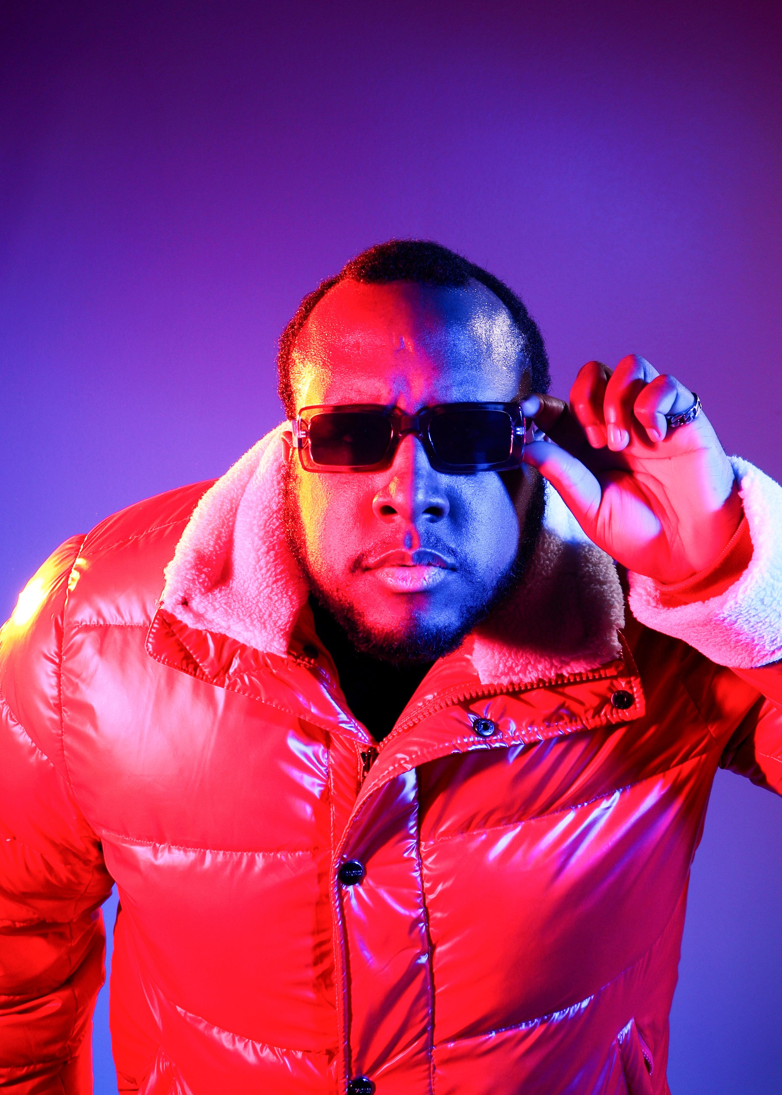 Fashion portrait of a man in a red puffer jacket with sunglasses against a vibrant purple background.