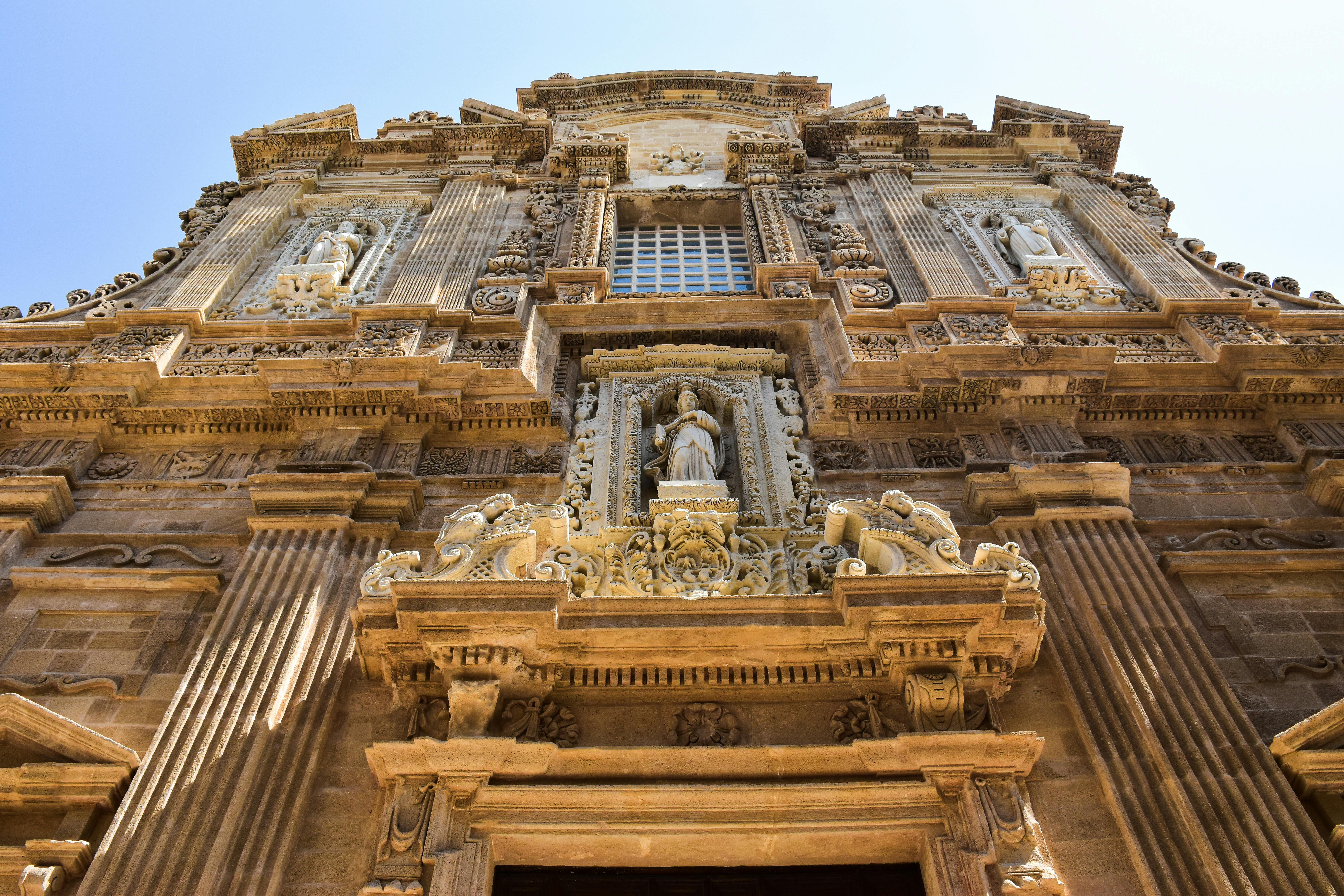 Sun-drenched Baroque facade of Santa Croce Basilica in Lecce, Italy, with intricate limestone carvings and warm golden light at mid-morning