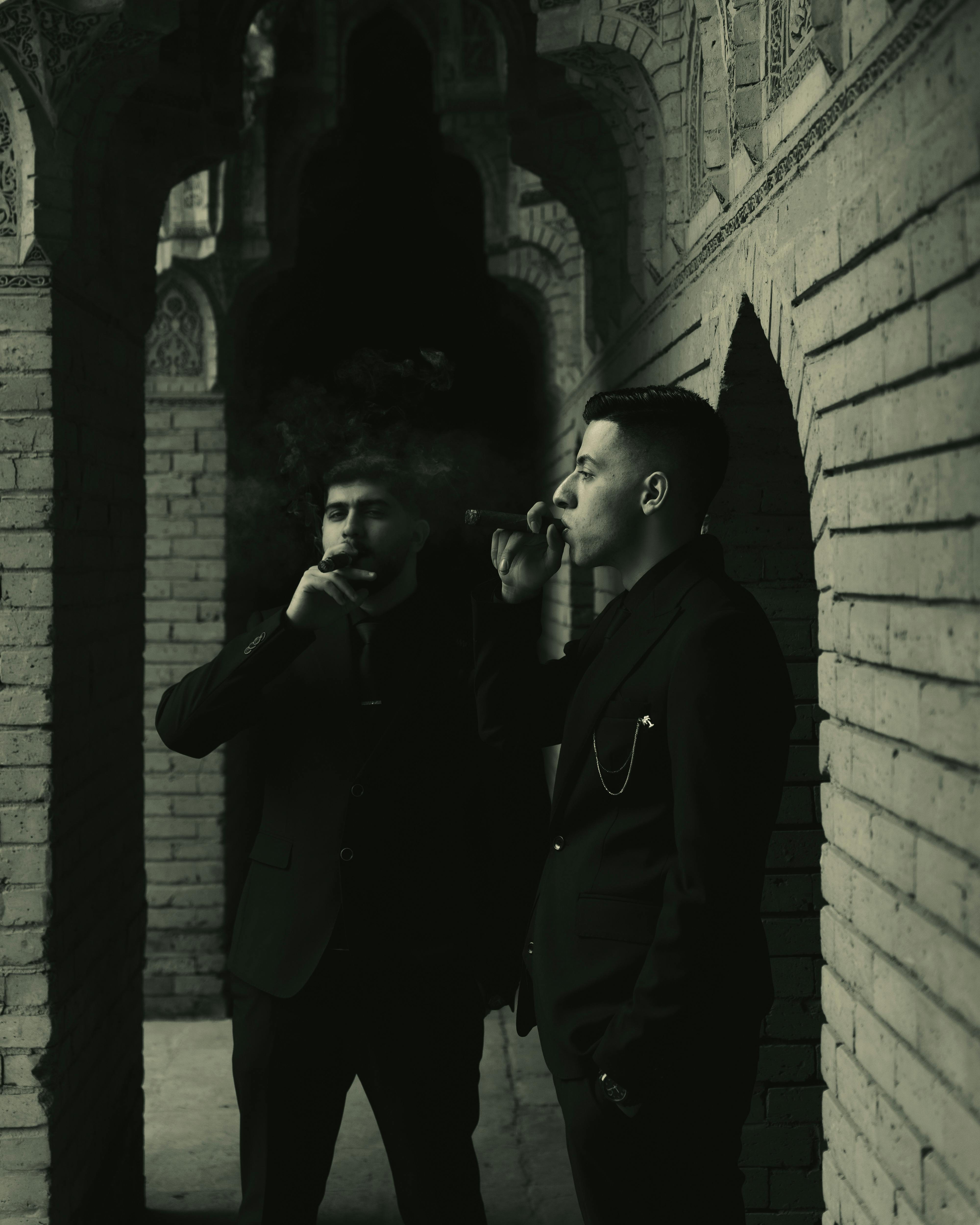 Two men in suits smoking cigars in a brick-arch corridor. Low-key lighting and atmospheric mood.