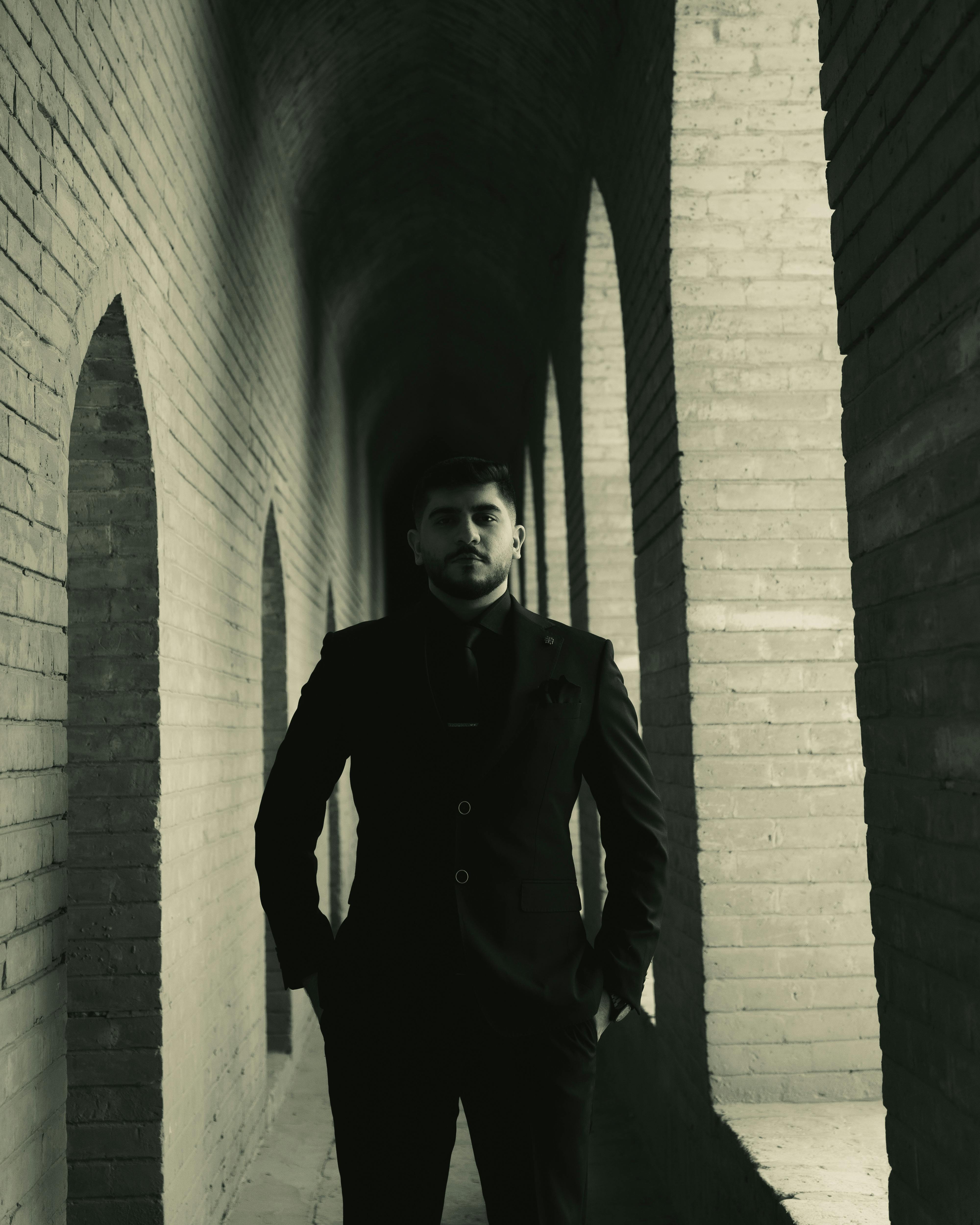 A moody portrait of a man standing in a historic brick corridor with shadows.