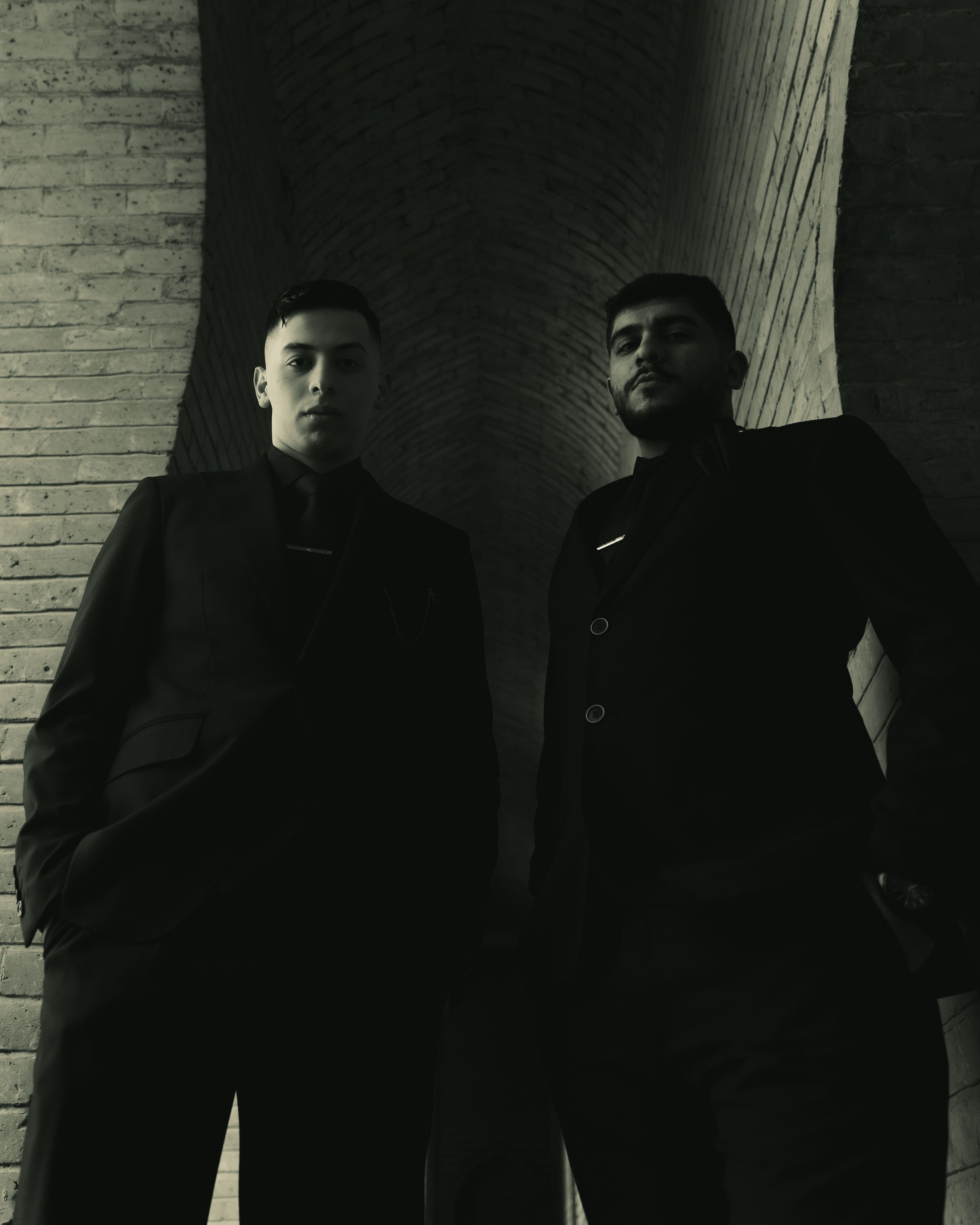 Two men in dark suits stand confidently under a striking brick archway.