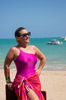 Smiling woman in a pink swimsuit relaxing by the beach under a clear blue sky.