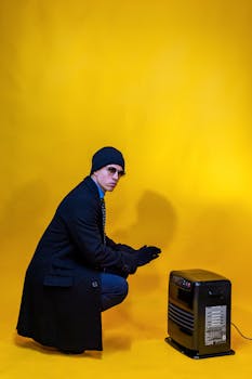 A man in winter attire crouches by heater against a vibrant yellow backdrop.