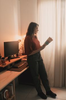 A woman enjoys reading near a window in a warm, cozy home office environment.