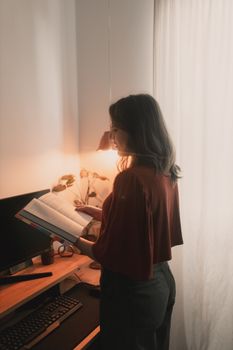 A woman stands by a desk, reading a book under warm lamp lighting.