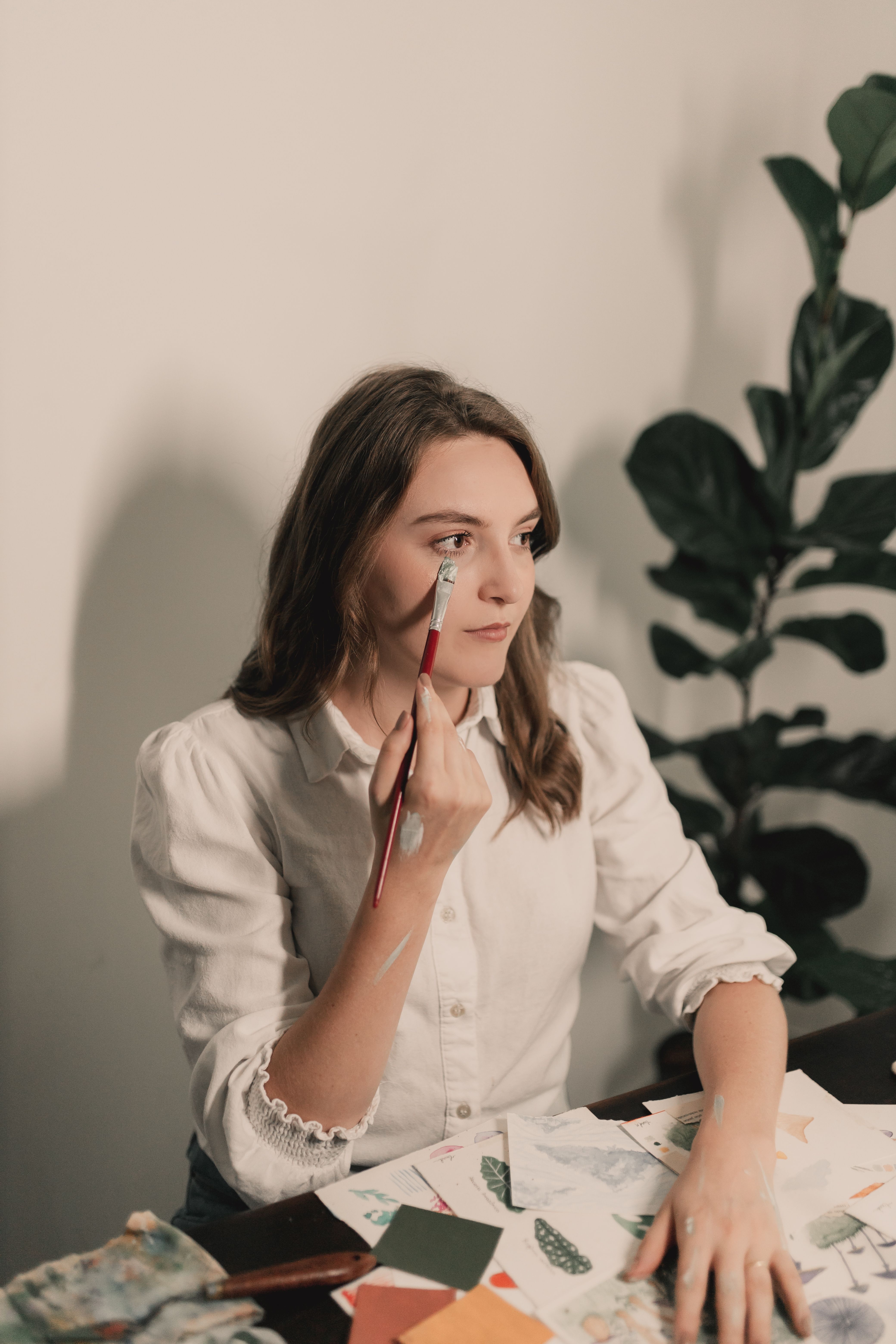 Creative portrait of a woman painting her face while seated at a table with art supplies.