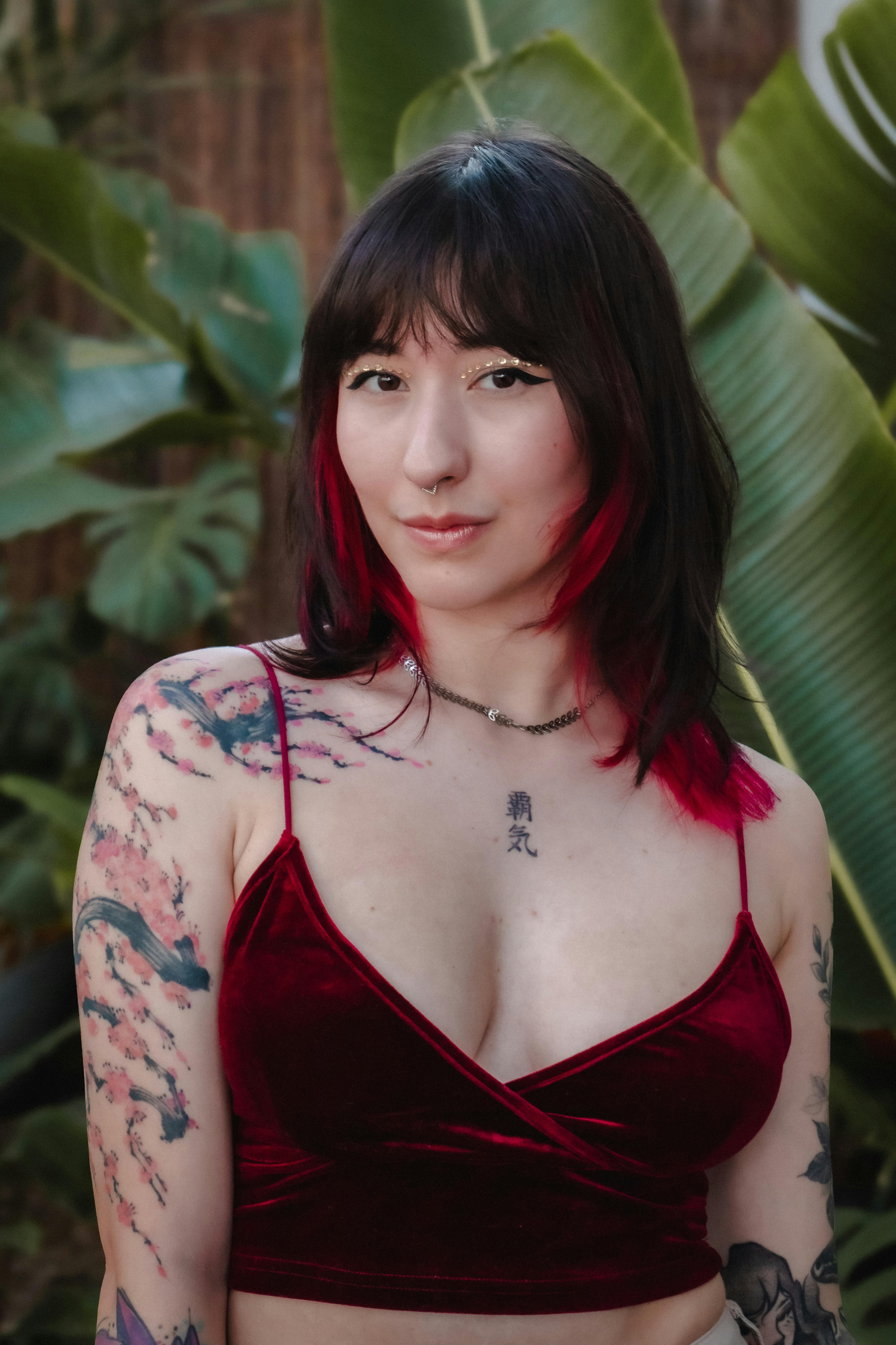 Woman with tattoos and red highlights posing in front of tropical plants.