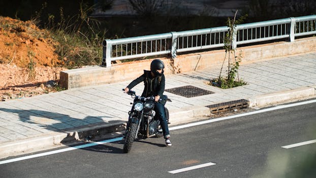 A biker with a helmet cruising on a motorcycle along an urban roadside curb under clear sunlight.