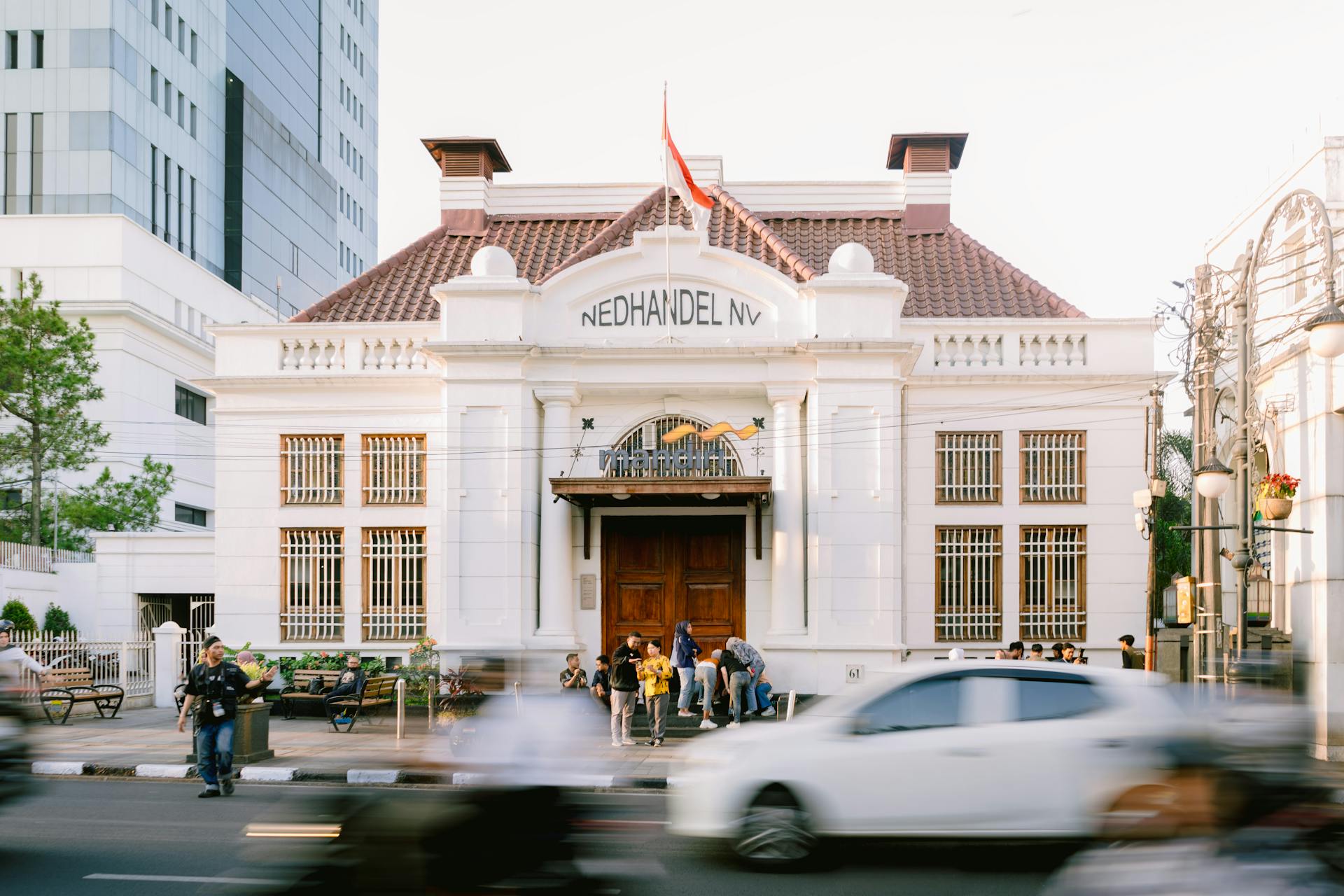 A lively street scene featuring the historic NED Handel NV building with blurred traffic motion.