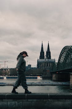 A young woman walks alongside the Rhine River with Cologne Cathedral in the background.