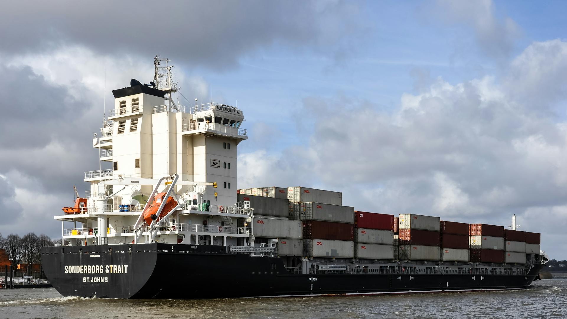 Container ship navigating Hamburg harbor with stacked cargo under a cloudy sky.