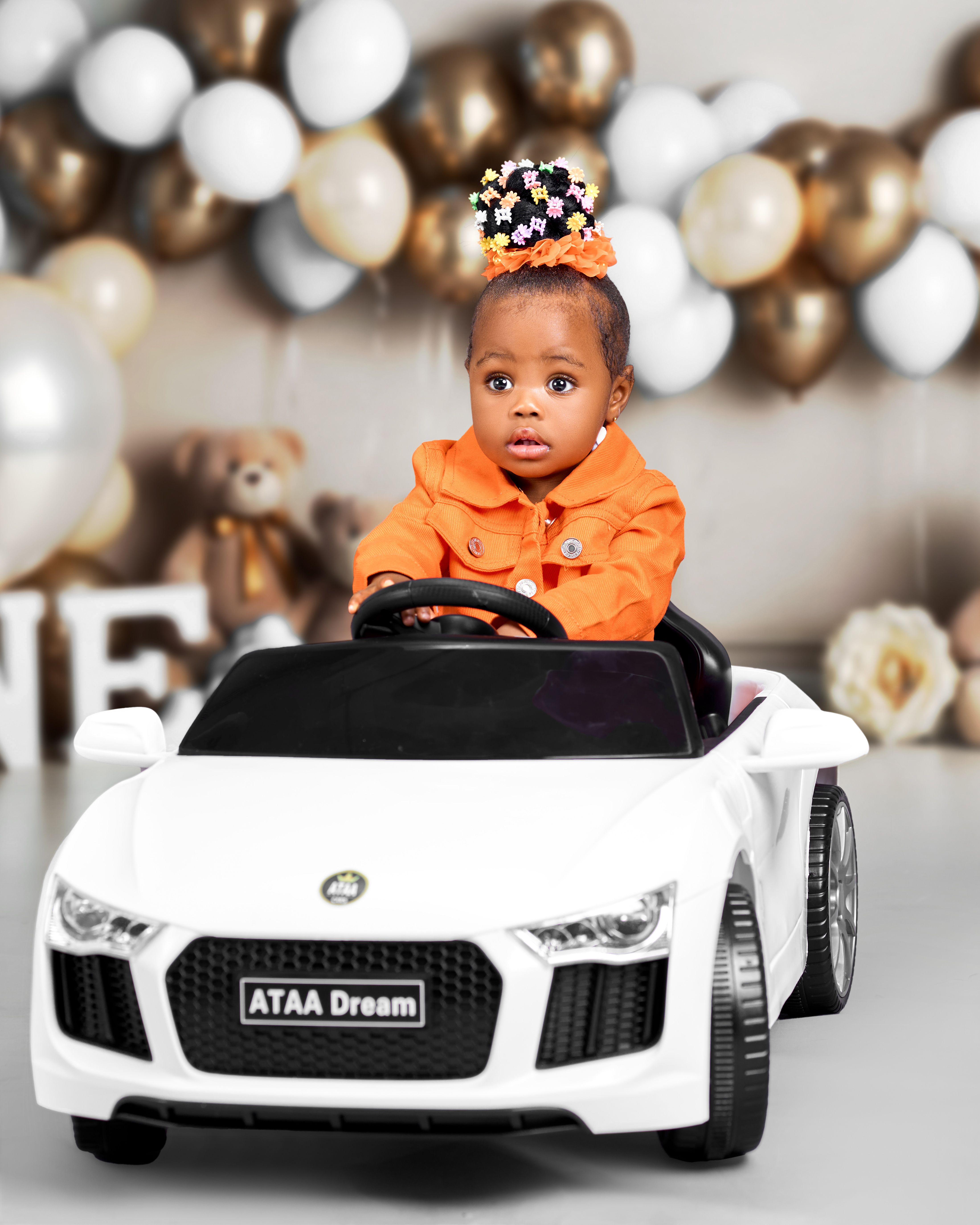 Charming baby in orange outfit driving a white toy car, surrounded by festive balloons.
