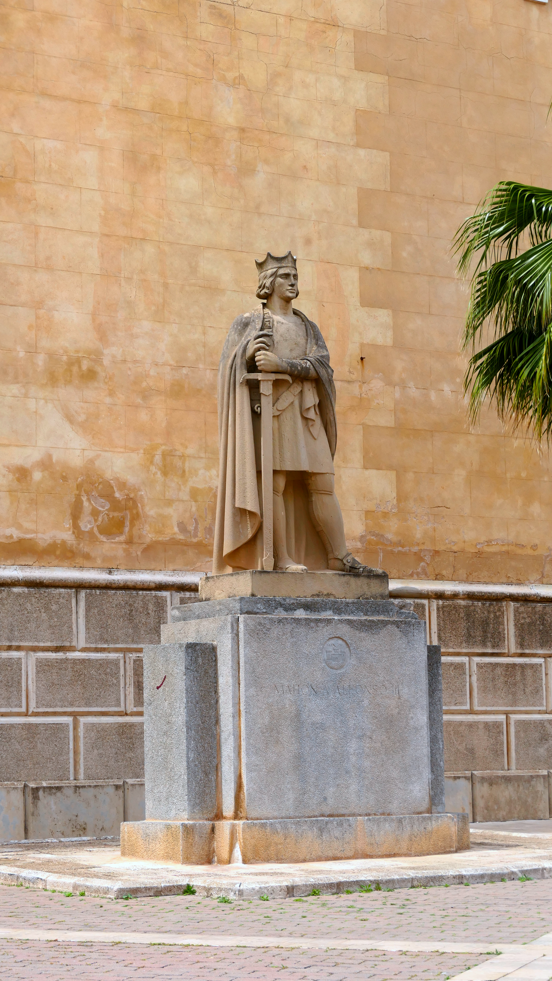 Statue of a medieval figure in Mahón, Spain, against a rustic wall.