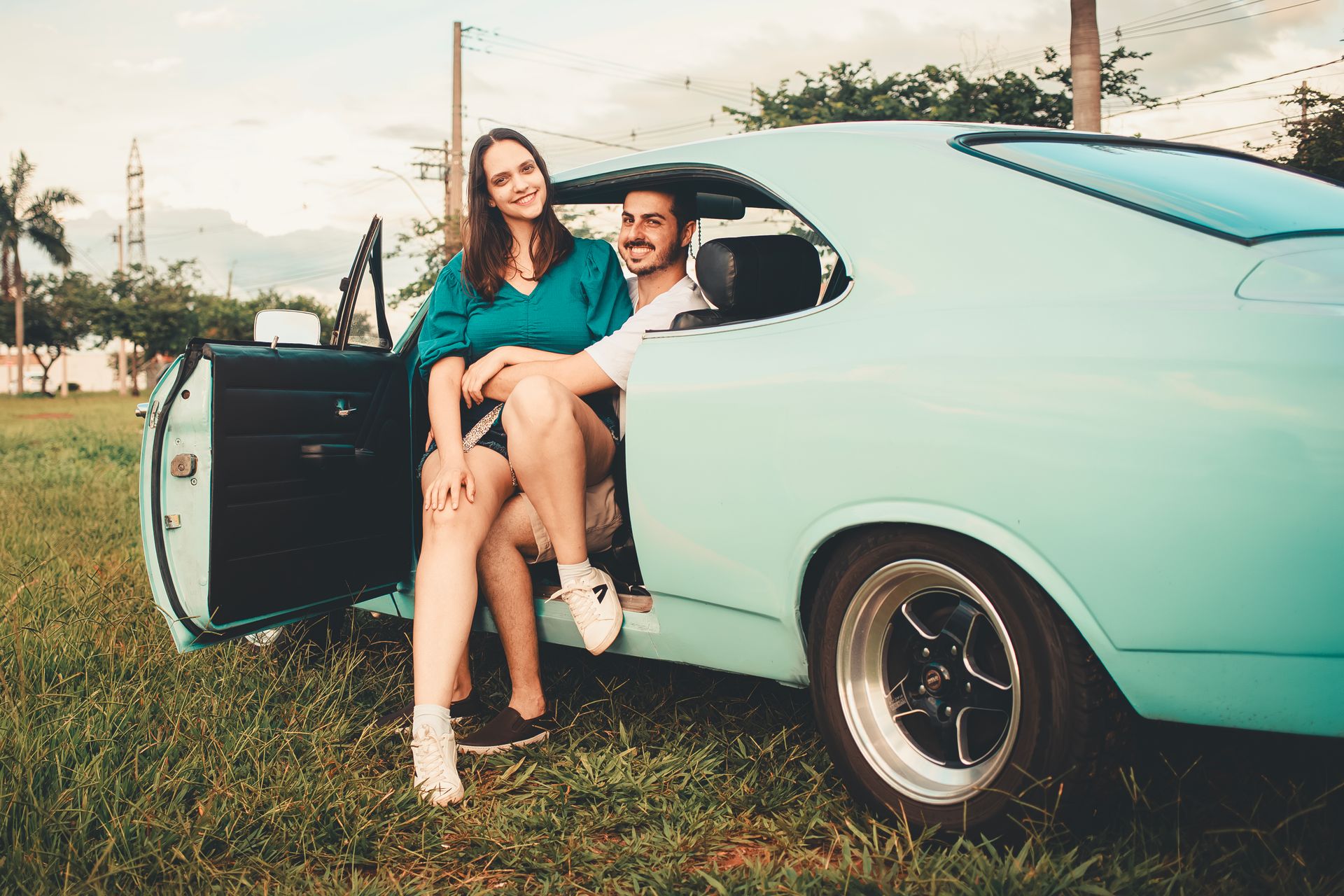 A happy couple enjoying the day sitting in a teal vintage car on a scenic grassy field.