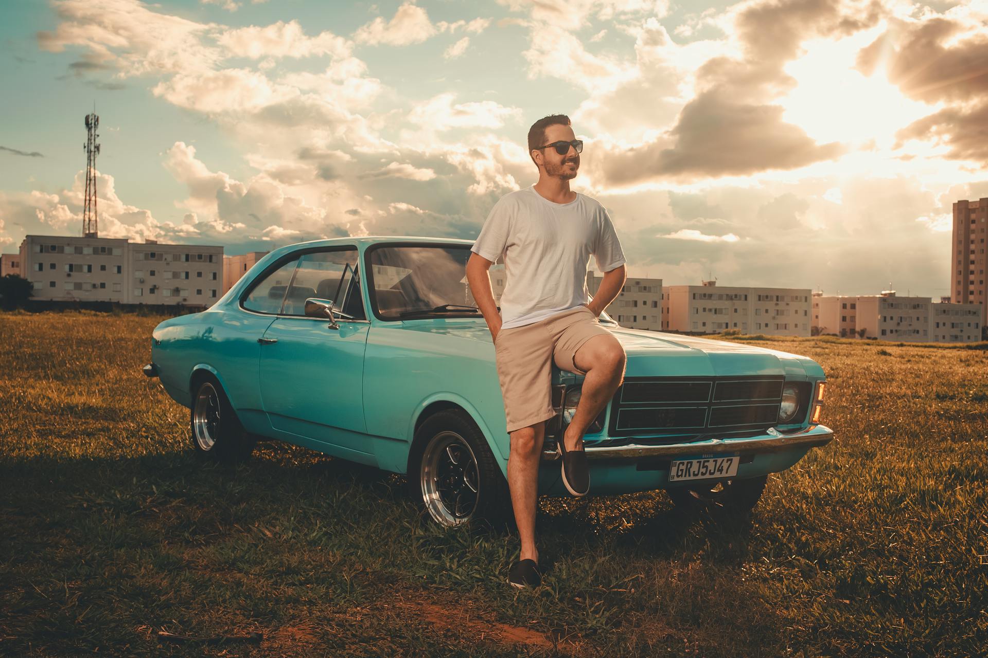 A man leans against a turquoise vintage car in an open field under a dramatic sunset sky.