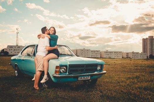 A couple embraces on a classic blue car in an open field with sunset skies.