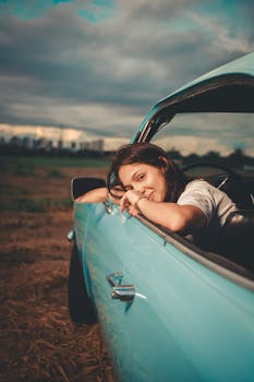 A smiling young woman leans out of a vintage blue car window during a scenic drive.
