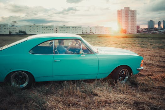 A classic teal car driven by an adult male on a field at sunset, city skyline in background.