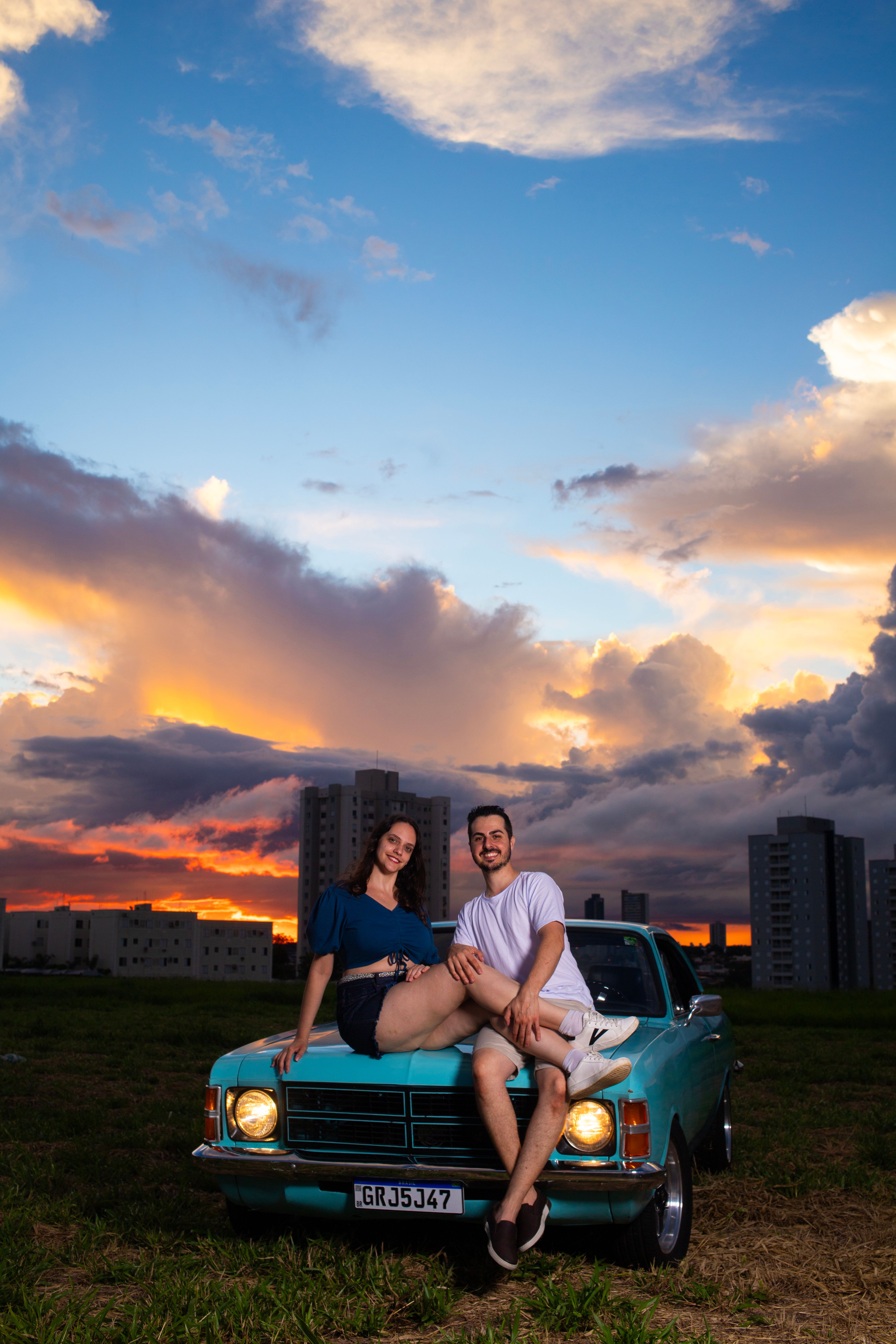 A couple enjoys a vibrant sunset atop a classic car with city skyscrapers in the backdrop.