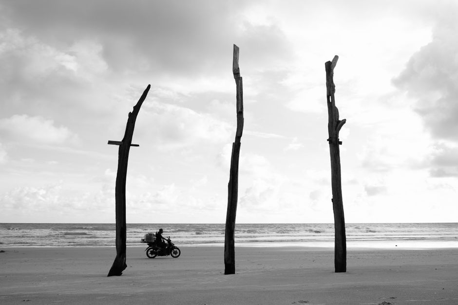 Black and white photo of a motorcycle by driftwood on a beach under cloudy skies.