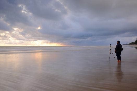 A person stands with a camera on a tripod by the ocean, capturing the dramatic sunset.