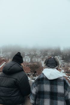 Two individuals in winter clothing observe a misty cityscape from above.