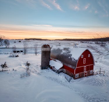 A scenic winter sunrise over a snow-covered red barn and silo in rural Minnesota.